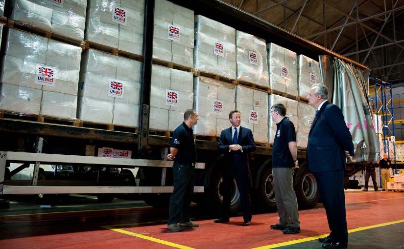 UK PM David Cameron visits a 'UK AID' facility in Wiltshire. He stands in a warehouse with boxes marked UK AID.