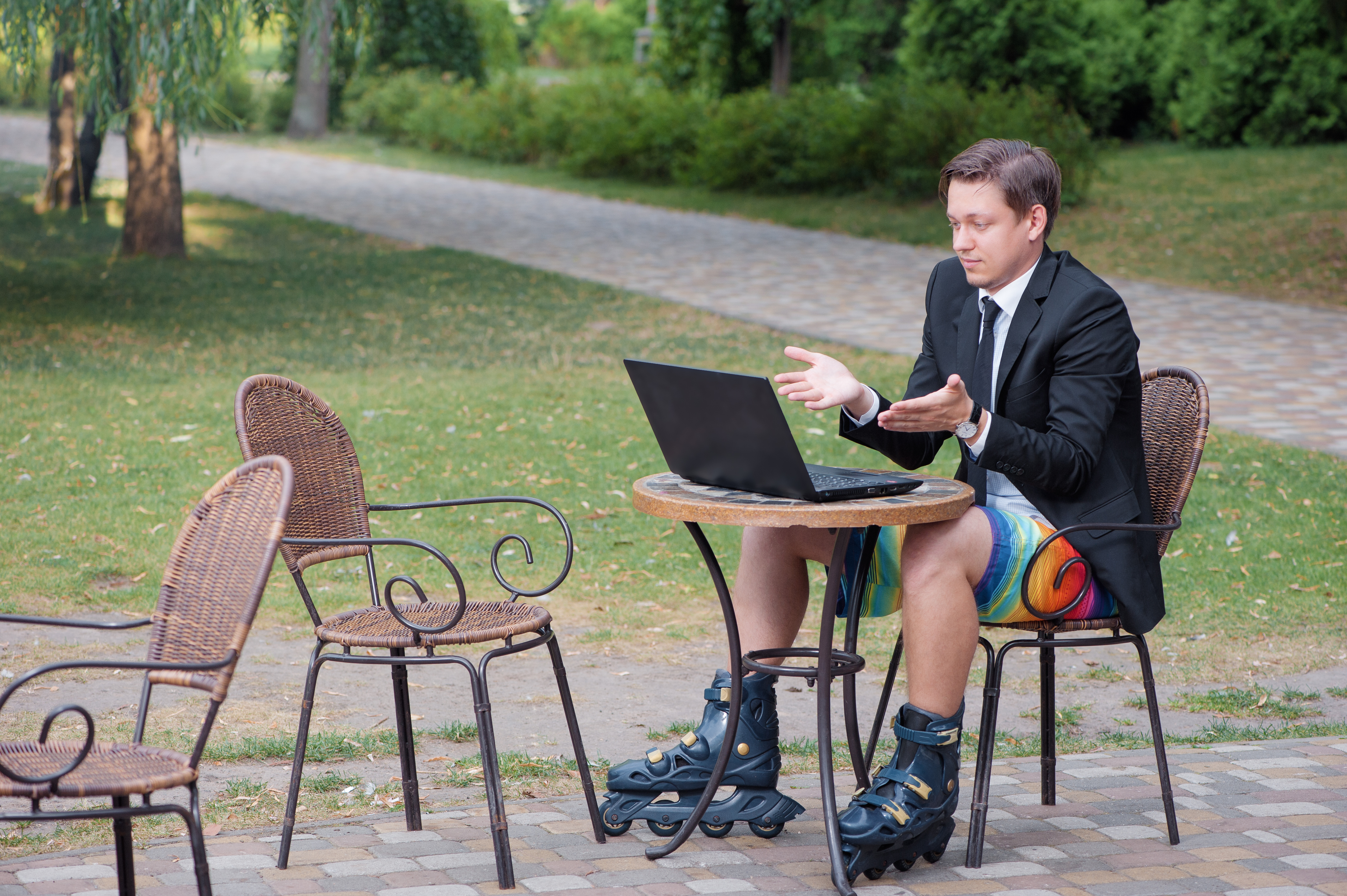 Businessman dressed in suit and shorts working with laptop, talking by skype at the park cafe outdoors, wearing roller blades